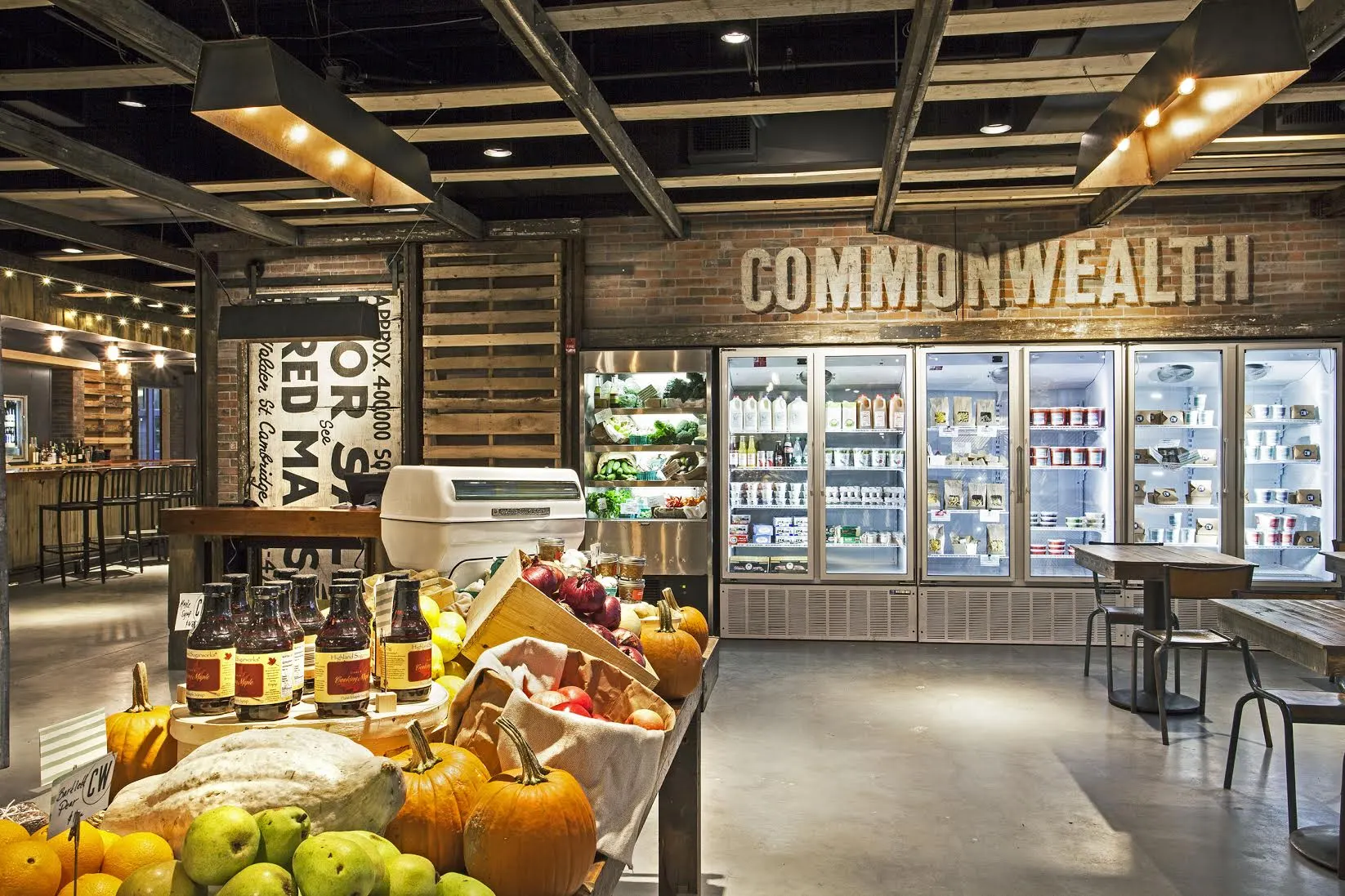 Commonwealth: a market counter of fruit and produce in front of refrigerated cases, beneath a Commonwealth sign on a timber wall