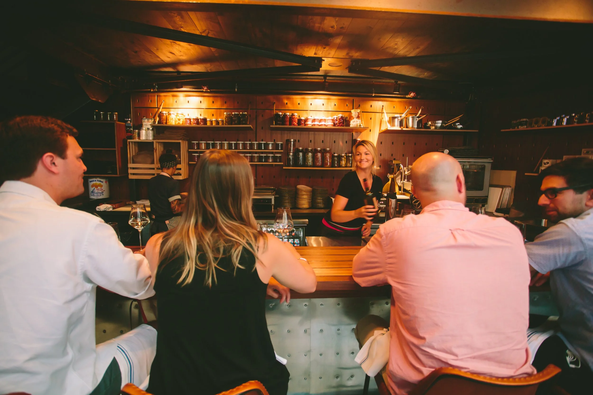Guests at the bar, with the bartender in front of shelves of tins and jars lit from above.