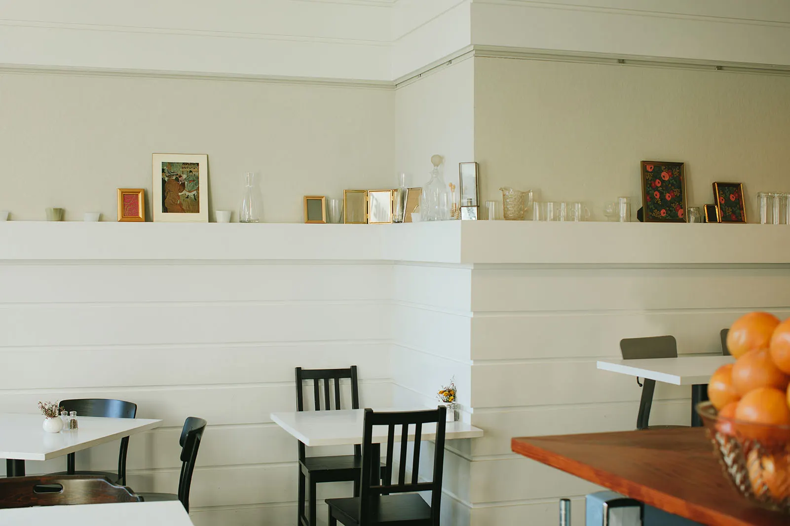 Small tables along a white-panelled wall, with a ledge of framed pictures and a bowl of oranges.