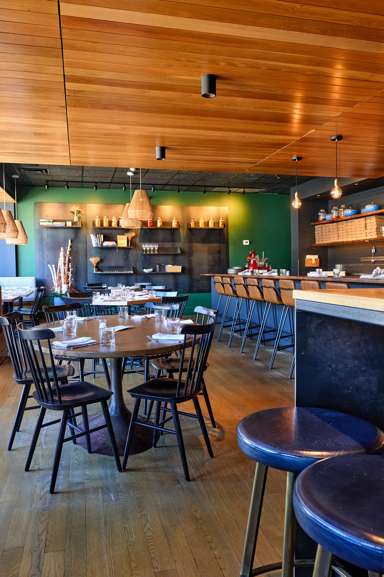 Round timber tables and black spindle chairs under a timber ceiling, with the green shelving wall and the bar beyond.
