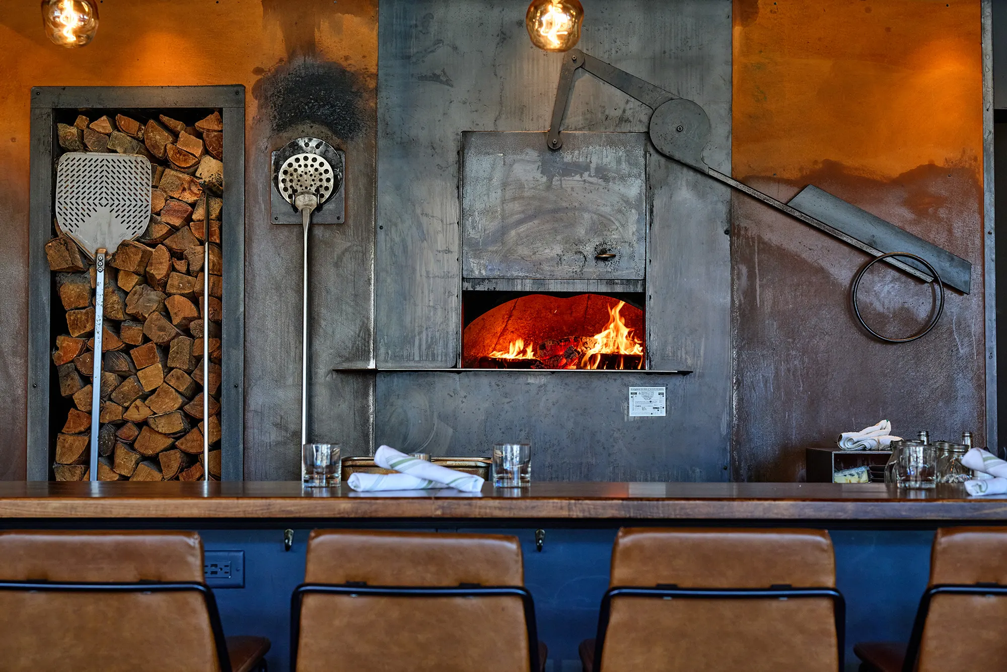 The steel-clad wood oven burning behind the counter, with stacked firewood, a pizza peel and a brush beside it.