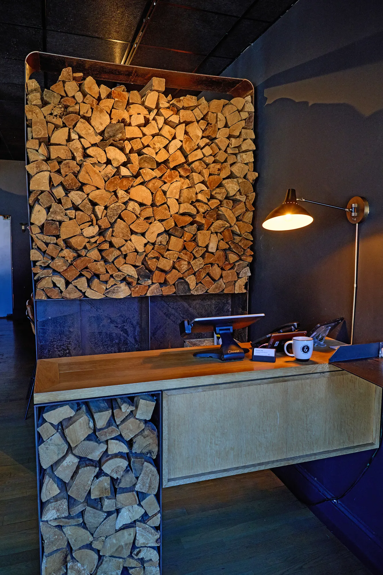 A host stand of pale timber over stacked firewood, lit by a wall lamp.