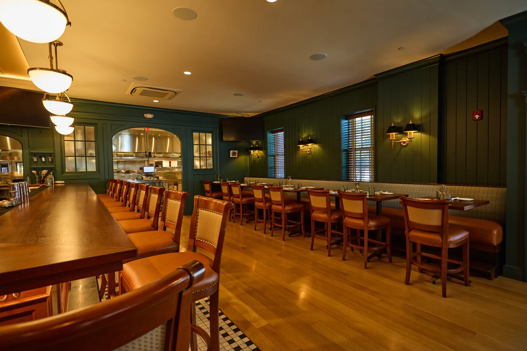 The long room at Puritan Tavern: a timber bar with leather stools on one side, tables along a plaid banquette on the other, green panelled walls, and the kitchen through an arched window at the end