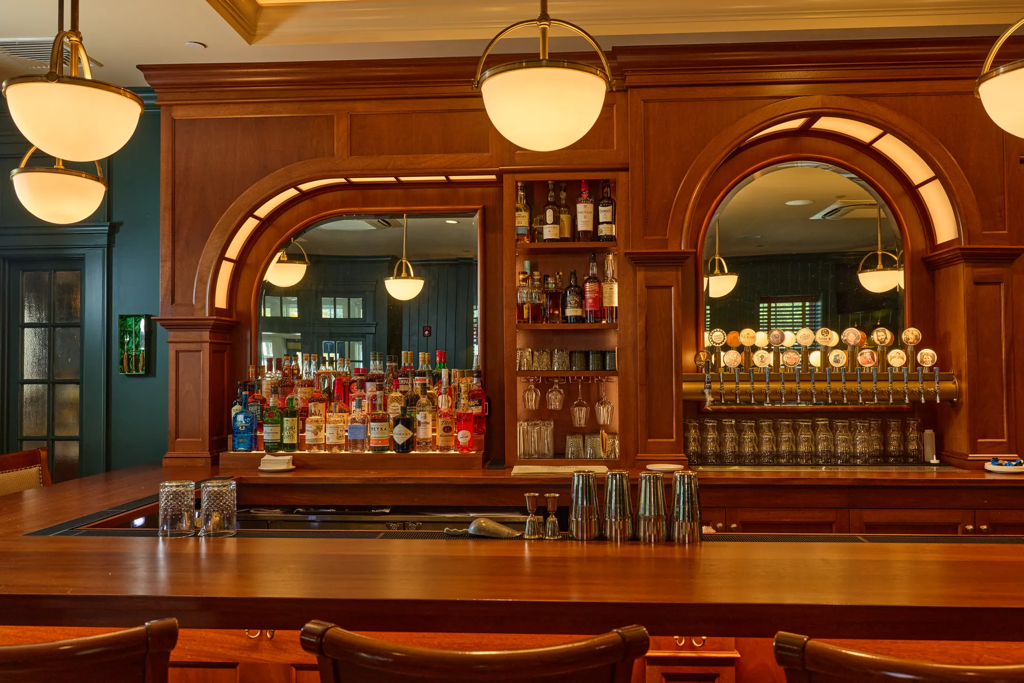 A mahogany back bar with two arched mirrors framing rows of brass taps, shelves of bottles between them, under globe pendants.