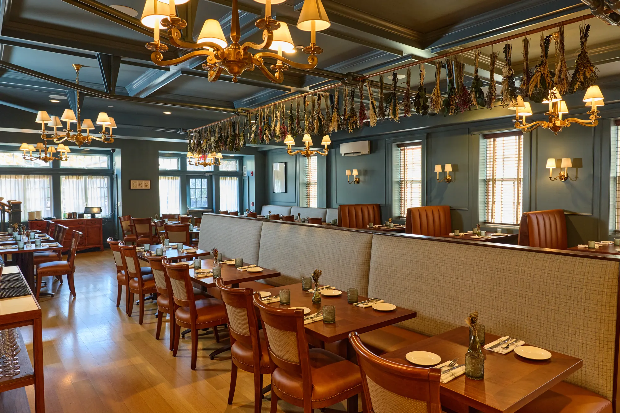 Dried flowers hung along a beam of a coffered ceiling with brass chandeliers, above rows of tables and a plaid banquette running the length of the room.