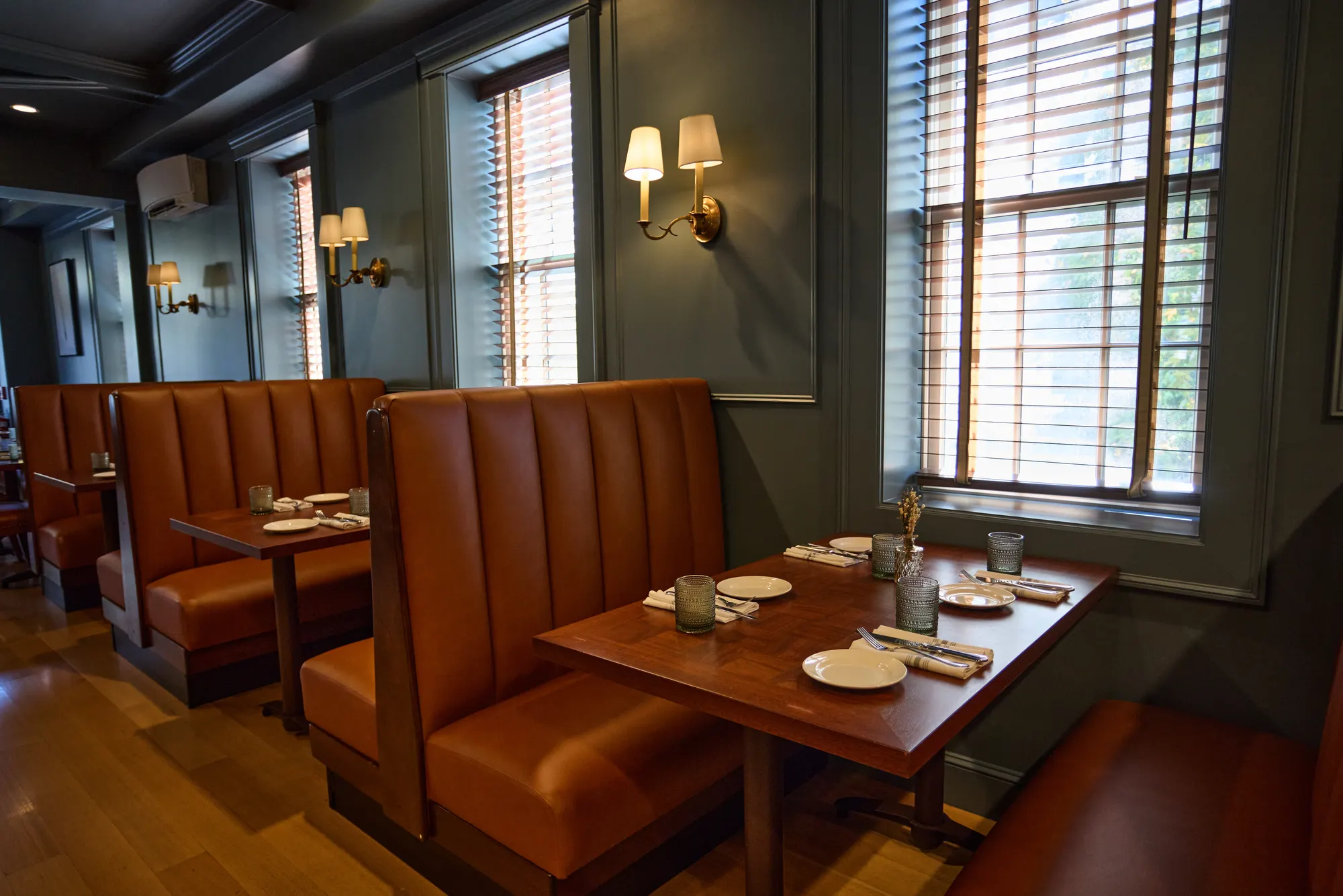 High-backed tan leather booths with set tables beside tall windows with timber blinds, brass sconces on the green walls between them.