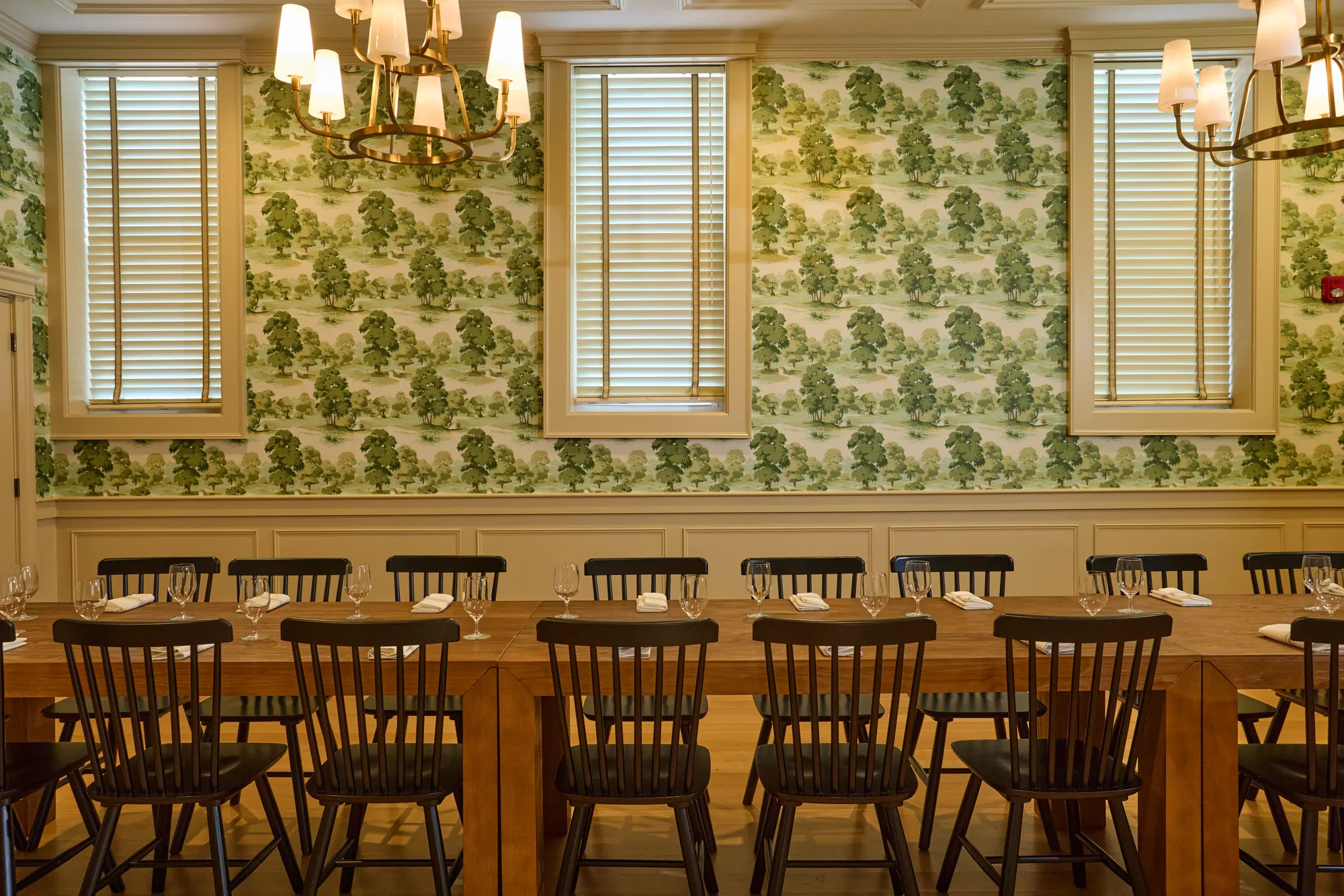 A long table set with glasses and black Windsor chairs against green toile wallpaper, between cream-framed windows, under brass chandeliers.