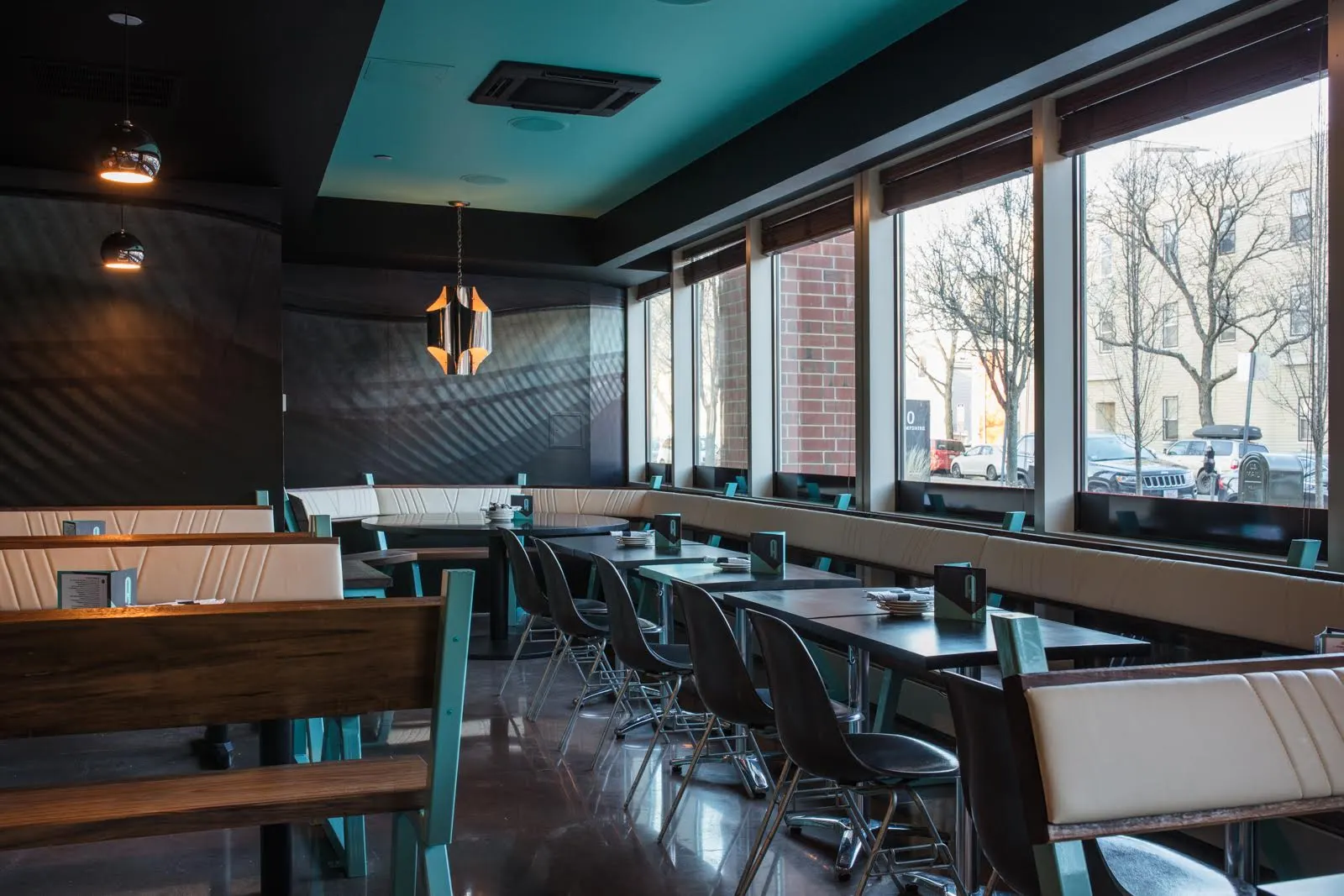 Booths and tables with teal-framed benches along a row of tall windows.