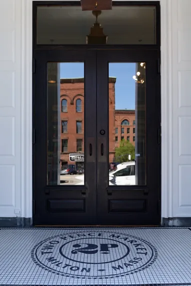 Glazed black double doors from inside, over a tiled floor with the Two Pence Market, Milton, Mass. seal in the mosaic.