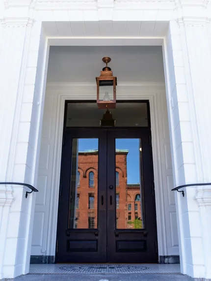 Tall black double doors in a white classical surround, with a lantern overhead and the brick building opposite reflected in the glass.