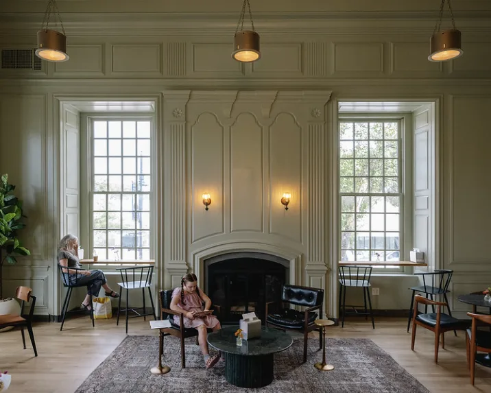 A panelled room with a fireplace between two tall windows, and people reading in armchairs.