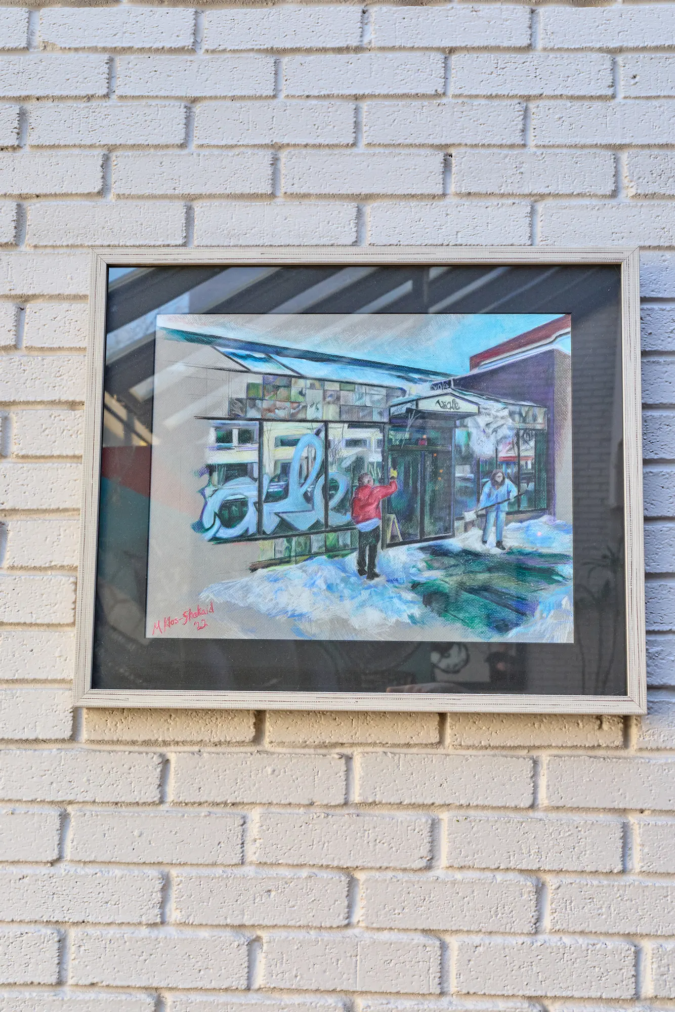 A framed painting of the street, hung on white brick.