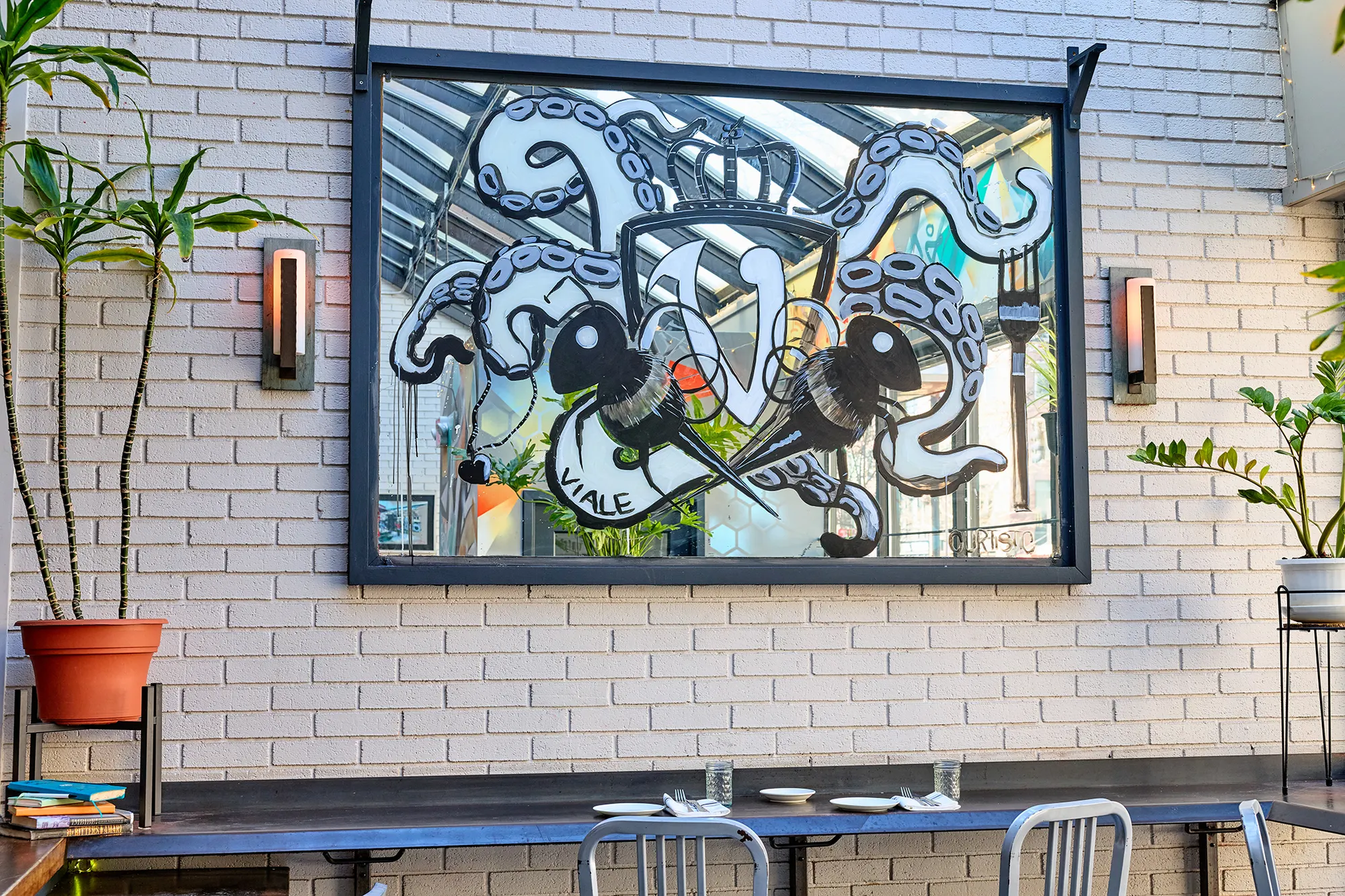 The framed black-and-white artwork on white brick, between plants.