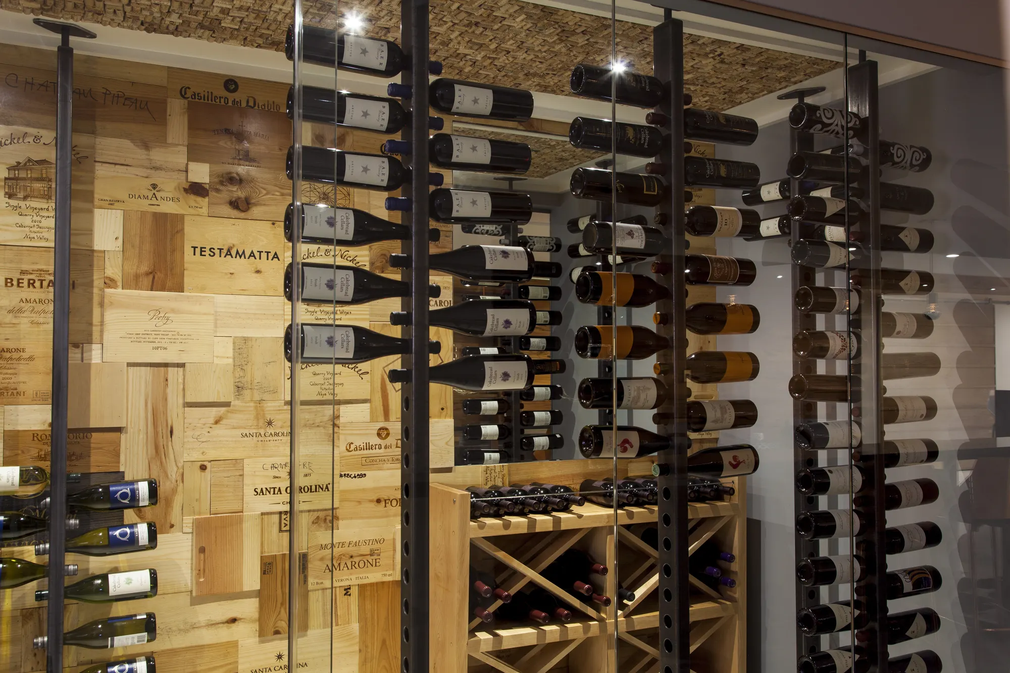 Bottles racked on steel rods against a wall of wine-crate panels, behind glass.