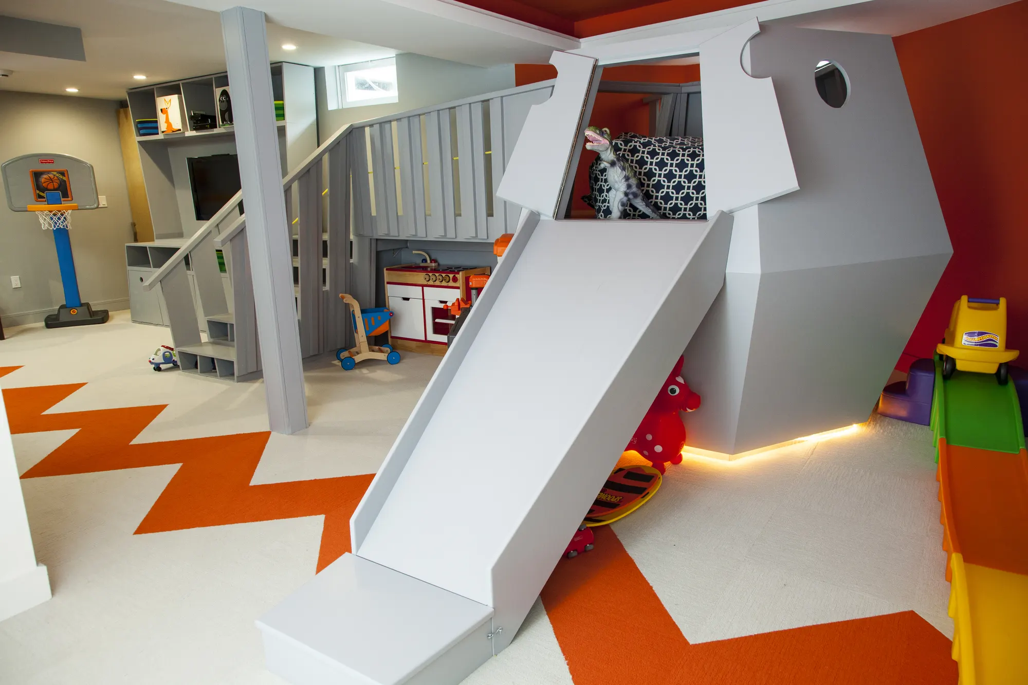 A grey slide coming down from a raised play structure onto a white floor striped in orange.