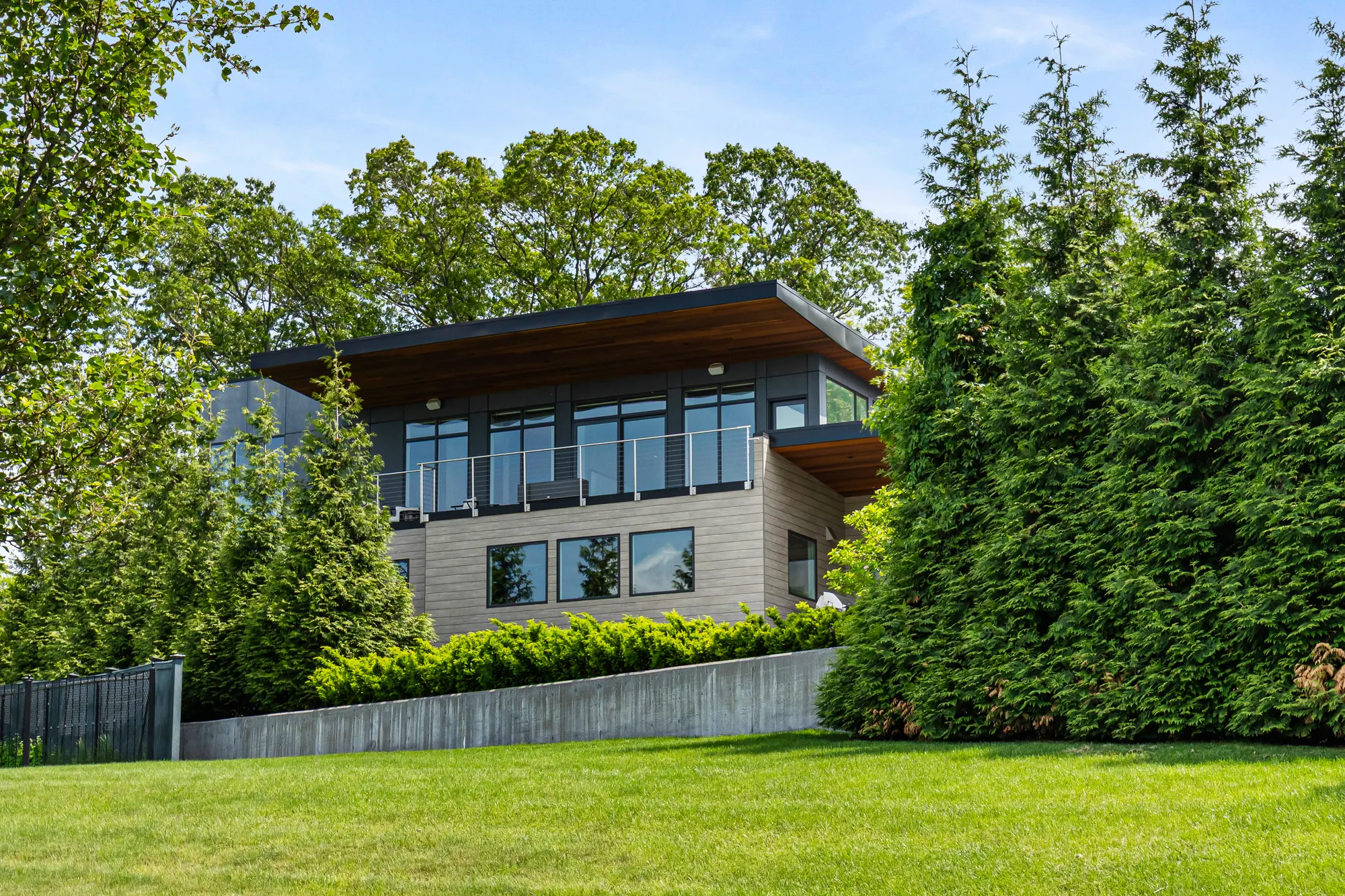 The house above a sloping lawn and a concrete retaining wall, between tall evergreens.