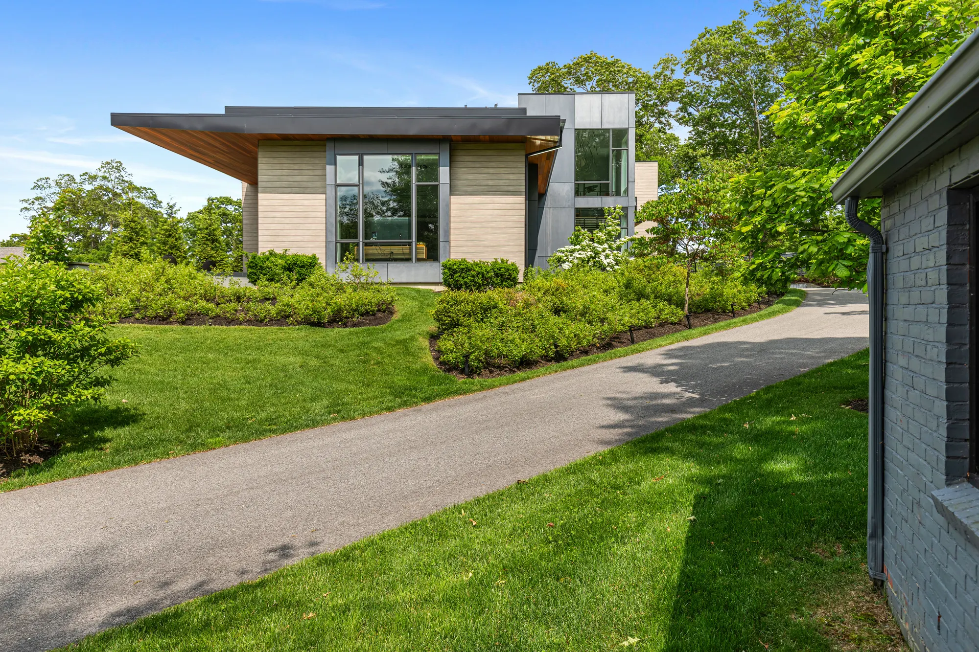 The driveway curving past planting toward the house, a corner of grey brick at the right.
