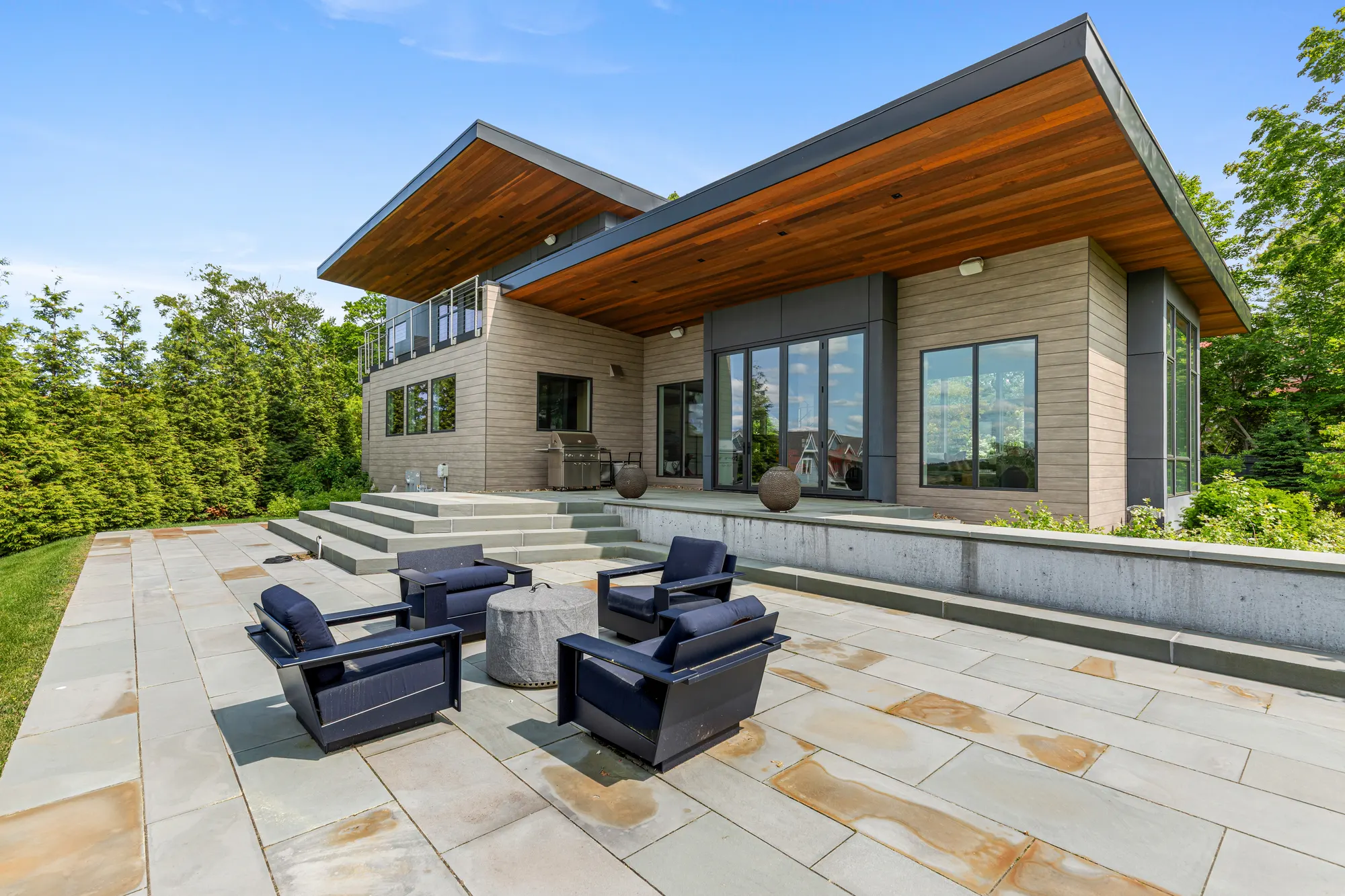 Four dark lounge chairs on a bluestone terrace, below the timber-lined overhang of the roof.
