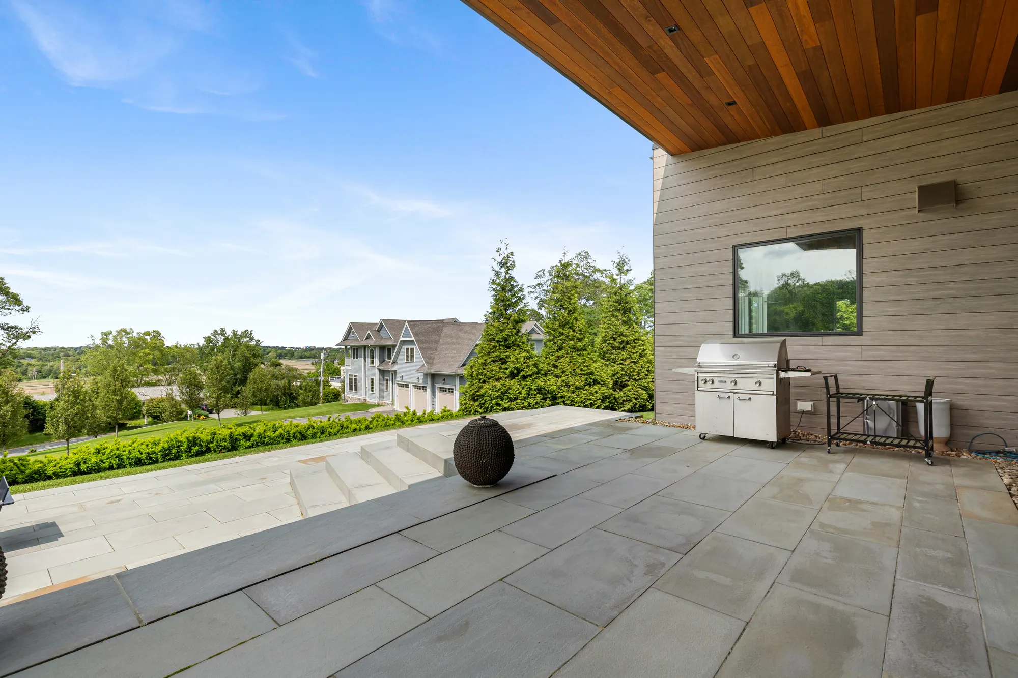 The terrace under the timber soffit, with a grill against the wall and a stone sphere on the paving.