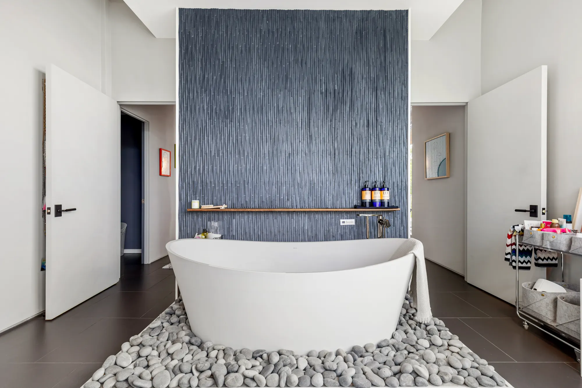 A freestanding white tub on a bed of river stones, against a textured dark blue wall.