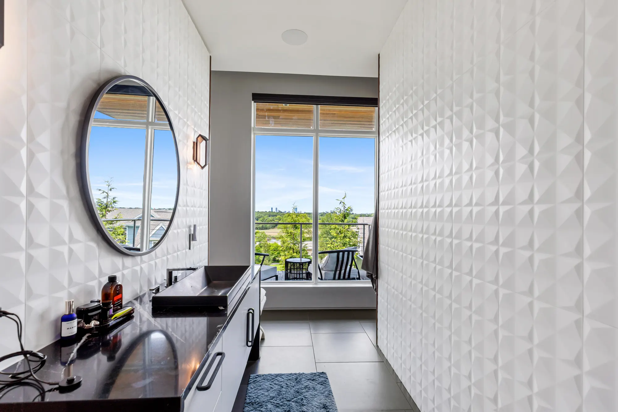 The white faceted tile wall and round mirror, with a window at the end of the room.