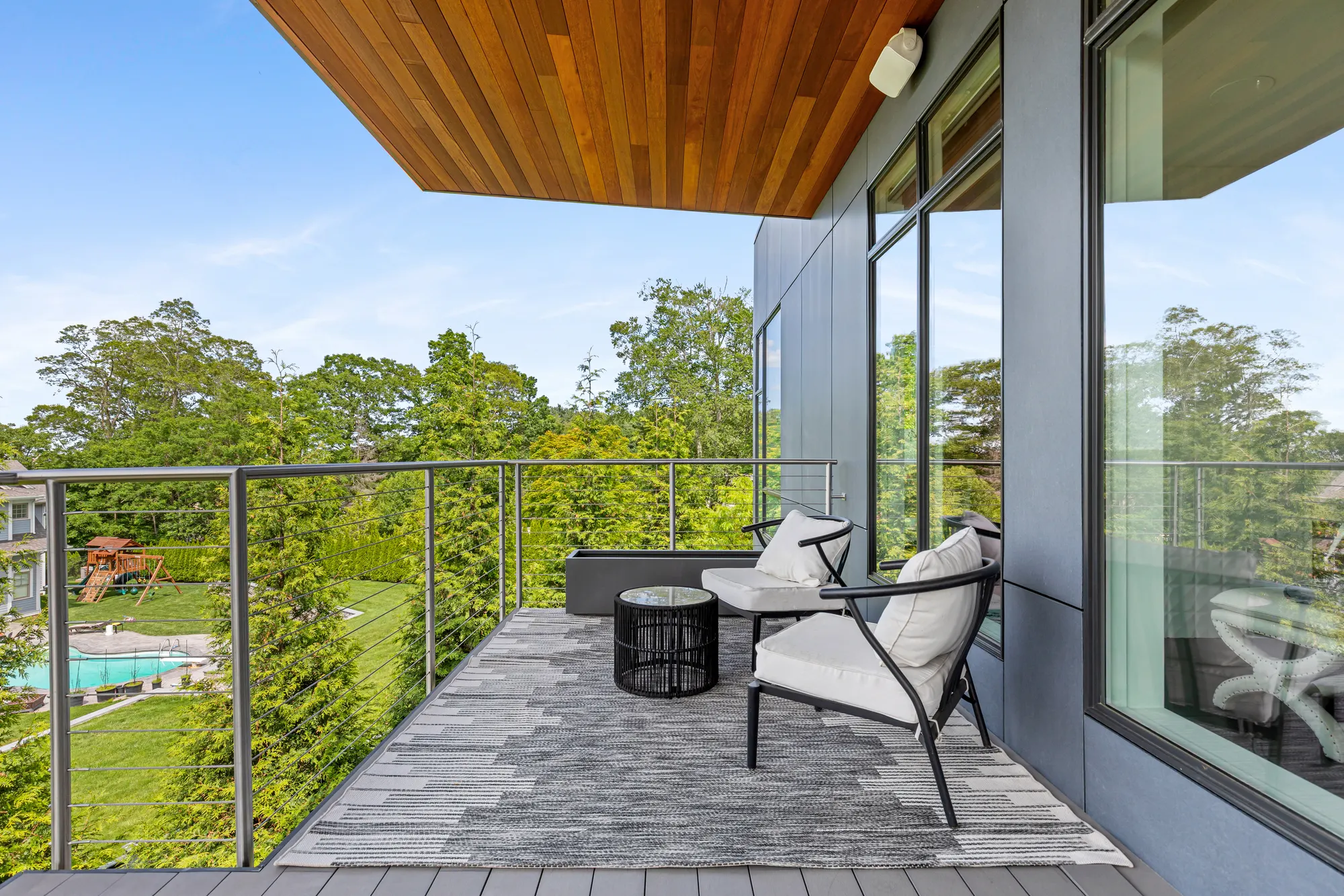 Two chairs and a side table on a rug on the deck, beside the glazing, with a pool below.