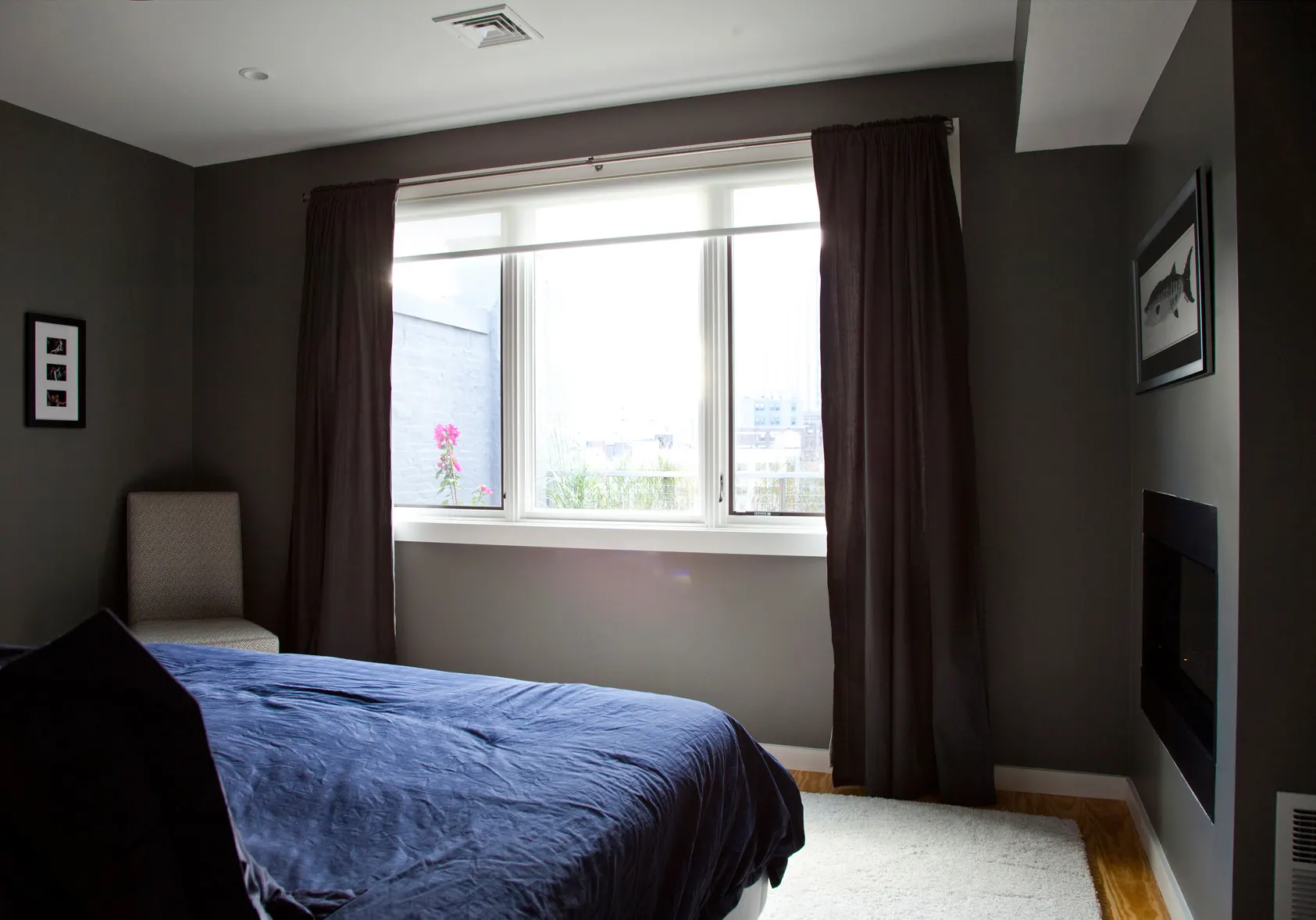 A bed under dark grey walls, with long curtains at the window and a fireplace set into the wall.