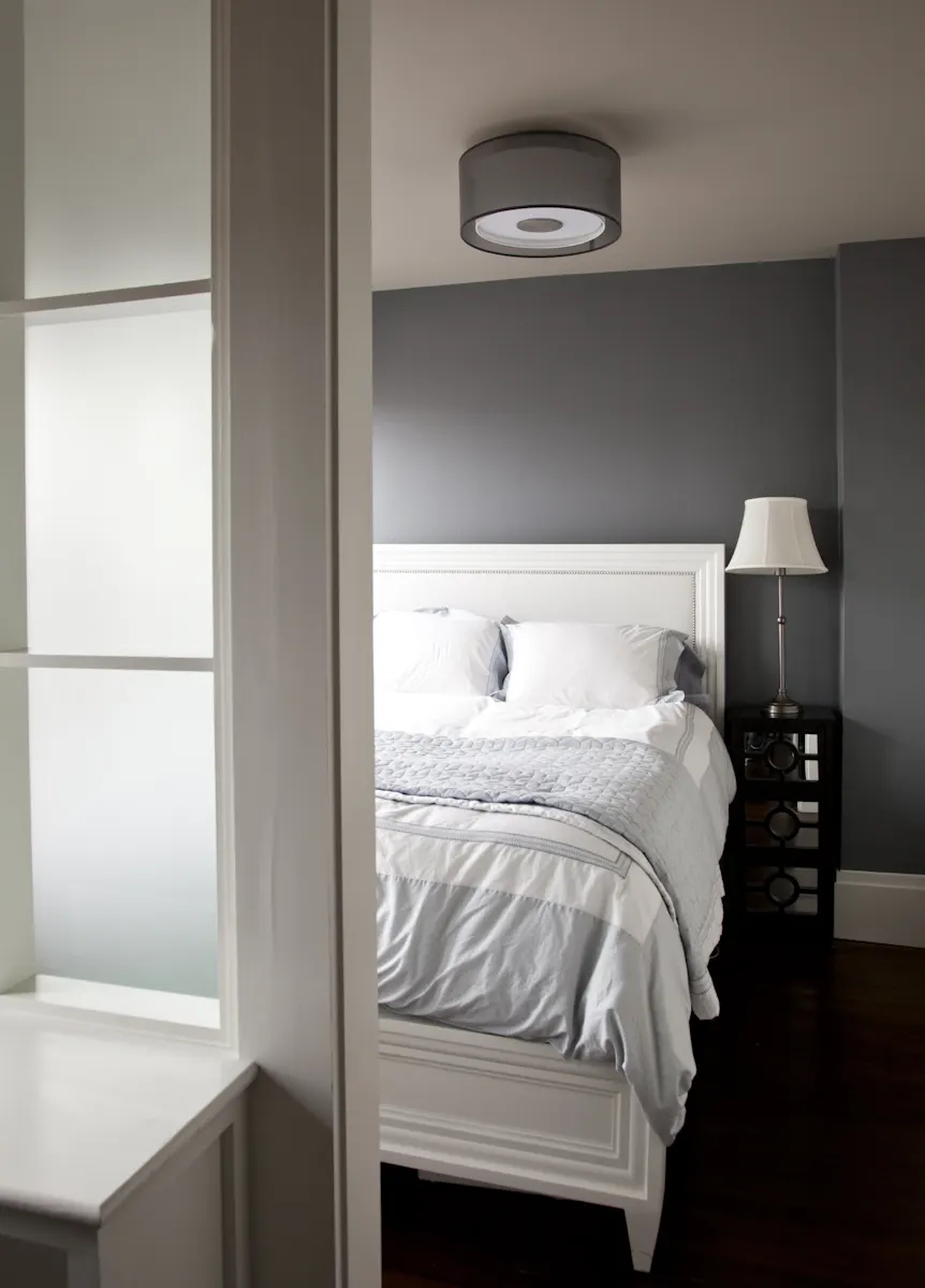 A white bed against a grey wall, seen past an interior window of frosted glass.