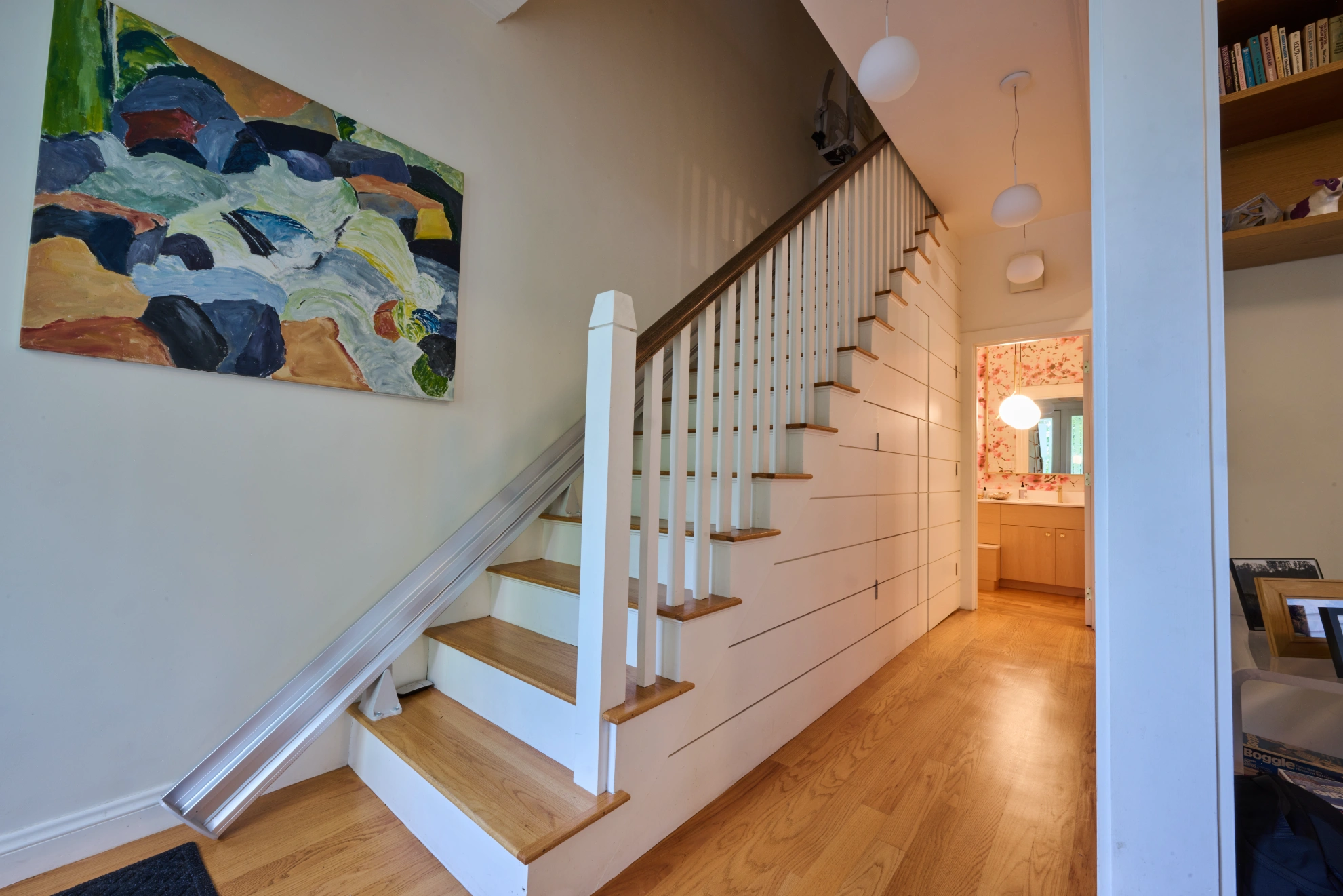 A white stair with a painted balustrade beside a boarded wall, with a painting above and the hall beyond.