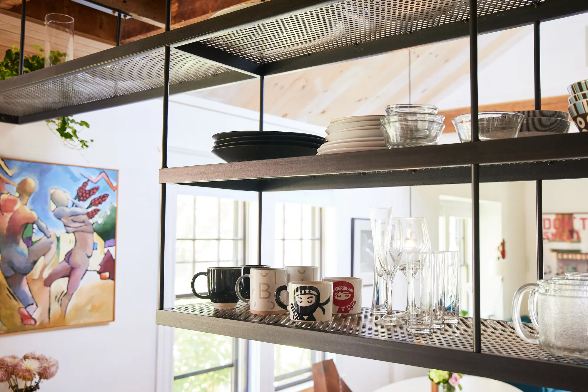 Mugs, glasses and plates on suspended steel shelves, with a painting behind.